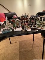 Wide view of five light-up village buildings arranged on table, including Toys for Tots House, Town Opera House, Dickens White Church, Ye Olde Bake Shoppe, and another building.
