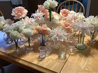 Photo showing more than 20 clear glass bud vases with a variety of silk flowers in soft white and blush pink tones arranged on a table.