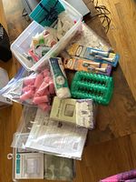 Photo of assorted bathroom supplies including pink hair curlers, foot massager, cleaning cloths, shower curtain, sheet snugs, lavender soap, and other items on wooden floor