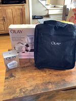 Photo showing the three main components of the lot: Olay Collagen Peptide Firming Duo box, additional Collagen Peptide 24 moisturizer box, and the Olay-branded black travel bag placed on a wooden table.