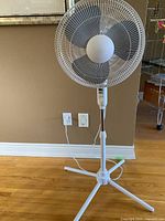 White oscillating floor fan standing on hardwood floor with power cord visible, showing full height and tripod base