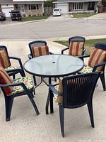 Full set displaying round patio table with six matching dark blue plastic chairs, each fitted with floral seat cushions and striped back cushions, arranged outside on a concrete surface.