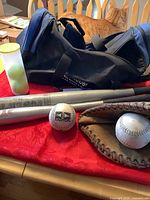 Blue and black duffle bag with baseball glove, two white softballs, and two aluminum bats displayed on red tablecloth.