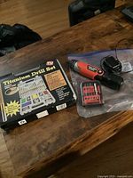 Black and Decker rechargeable screwdriver, charger, and red bit case alongside the new titanium drill set box on wooden surface.