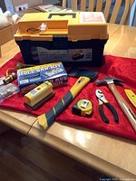 View of toolbox with tools arranged on a table showing hammer, cutting tool, axe, measuring tape, pliers, couplers, sanding block and hole saw kit box.