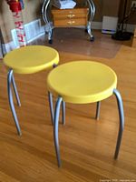 Two stools with yellow plastic round seats and metal legs on a hardwood floor, showing the top and side view of seats and legs.
