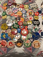Overhead shot of approximately 60 assorted vintage advertising pin-back buttons laid on quilted surface