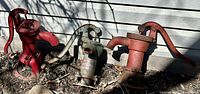 Three antique cast-iron hand pumps grouped outside against house siding, one painted red and two with green and redish patinas, showing overall condition and size comparison.