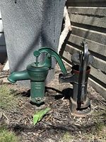 Photo of both antique water pumps placed near a garden wall, showing their size and appearance.