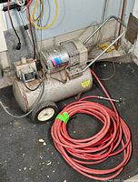 Full view of Campbell Hausfeld air compressor with wheels and handle, positioned on floor with coiled red hose next to it.