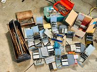 Overview photo showing a large collection of opened and closed drill bit index cases and a wooden box with long drill bits on a garage floor.