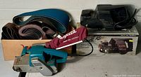 Makita belt sander with dust bag, second belt sander on original box, and box of new sanding belts