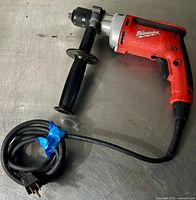 Milwaukee heavy duty corded drill with side handle and long power cord laid out on metallic surface.