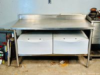 Front view of stainless steel table showing welded backsplash, lower shelf, and two loose metal drawers underneath.