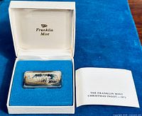 Front view of sterling silver ingot bar inside original white presentation box with blue velvet interior and certificate of authenticity booklet beside it.