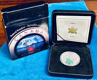 Coin in black velvet-lined display box with certificate of authenticity and outer packaging visible on blue background.