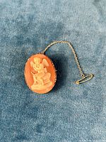 Front view of oval coral cameo brooch pendant with cherub figure and safety chain laid on blue fabric.