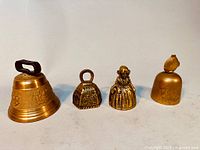 Four antique brass bells displayed: a large Swiss bell with a wrought iron handle, two small figurative female brass bells, and one Chinese fish-shaped brass bell.