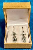 Pair of sterling silver dangle earrings with clear cubic zirconia stones presented inside a beige jewelry box with white cushioned interior.