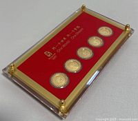 Five gold-tone medallion coins arranged horizontally in clear round mounts on red backing inside a rectangular clear acrylic case with gold corner accents. Chinese text and 'One World One Dream' slogan visible.