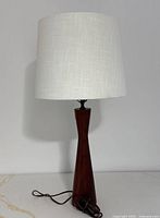 Front view of the complete table lamp with white textured fabric shade attached, showing the conical solid teak wood base and electrical cord.