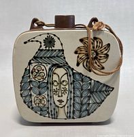 Front view of the flask showing the stylized face and decorative floral and leaf patterns in muted blue, black and tan on a cream background.