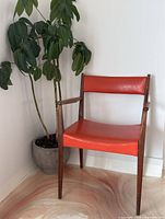 Side and angled view of the teak armchair showing red vinyl seat and backrest against a wall and plant.