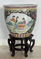 Full view of the ceramic planter on its wooden stand showing painted traditional Asian scene with people and koi fish designs.