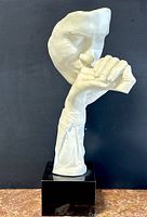 Front and side views of signed white sculpture depicting a face resting on a clenched fist on black base
