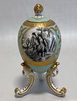 Front view showing black and white pastoral scene of a couple and gold accented turquoise body of egg on ornate tripod stand.