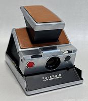 Polaroid SX-70 Land Camera unfolded showing lens, red exposure button, adjustment controls, and front panel labeled 'POLAROID SX-70 LAND CAMERA'.
