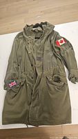 Olive green 3/4 length men's jacket laid flat, showing Canadian flag patch on right sleeve and Union Jack patch on left cuff.