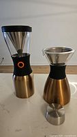 Both insulated coffee makers shown side by side on a white counter; stainless steel bases with copper finish and black silicone grips visible.