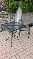Black metal mesh garden table and chair set on brick patio with stone wall background