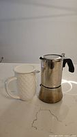 Photo shows the full espresso maker alongside the white ceramic tea mug with lid, placed on a kitchen countertop.