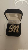 Silver tone 'M' brooch in black jewelry box showing front side with cursive style letter 'M'.