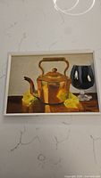 Frontal photo of the oil painting showing a copper teapot, glass, and yellow cloth in a light frame, signed by artist.
