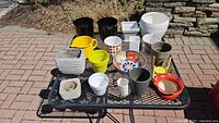 Overview of the 22 assorted planters and pots including various colors and materials seen on a black metal outdoor table on a stone and brick patio.