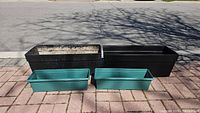 Four plastic window boxes seen outdoors on a brick surface, two large black boxes at the back and two smaller green boxes in front.