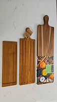Three wooden kitchen/charcuterie boards laid out on a white surface: left smooth narrow bamboo board, middle bamboo grooved board, right large acacia paddle board with handle.