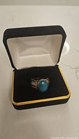 Ring shown inside a black and white jewelry box with gold trim. The ring has an oval blue stone set on a wide sterling silver band.