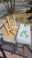 Two wooden plant stands, pine color, displayed in an open box outdoors on a black metal table.