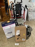 Two red and black walkers with seats and bags, box for Drive shower bench, additional packaged mobility aid, and walking boot visible on carpeted floor next to floral couch.