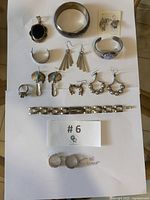Top view of all 12 pieces laying on white paper, showing varied designs and mounting, including earrings, rings, pendants, bracelets, with one pair of earrings noted as not stamped.