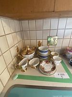 Wide shot of all 13 china pieces including sugar bowl, creamers, teacups, saucers, and dishes arranged on a kitchen counter with tiled backsplash.