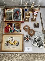 Overview photo showing vintage automotive magazines, collectible patches, multiple trophies, 1910 Hudson framed print, automotive plaques, chrome cap, cigarette tin, and black-and-white photos.