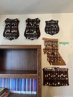 Five wooden wall mounted spoon display holders mounted on a wall, each holding multiple souvenir teaspoons. Includes a California-shaped holder.