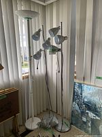 Five lamps in a corner including one white floor lamp with round top, two multi-headed floor lamps with gray adjustable heads, and two gray clamp desk lamps with flexible arms. Lamps are on a wooden floor with vertical blinds behind.