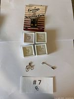 Seven jewelry pieces displayed including two pendant earrings with white opal stones, two pairs of stud earrings with clear stones, one pair of clip-on earrings for sensitive ears, and a small stick pin with clear stone.