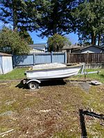 Side view of white fiberglass boat on galvanized single-axle trailer showing hull shape, seating wells and cover over bow